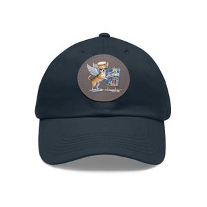 May include: Navy blue baseball cap with a circular patch featuring a chihuahua with angel wings and a halo, painting on a small easel. The chihuahua is brown and white, and the painting has bright colors. The cap has a curved brim.