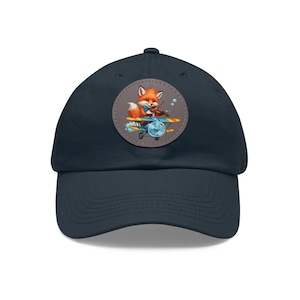 May include: A navy blue baseball cap with a round patch featuring a cartoon fox playing the violin while flying a small airplane. The fox is orange with a white chest and a blue bow tie.