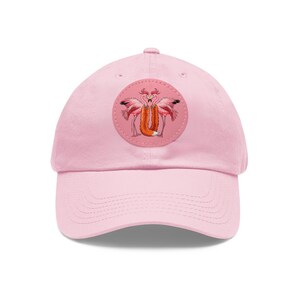 May include: A light pink baseball cap with a curved brim. The cap features a circular patch with a whimsical design of two pink flamingos embracing a fox. The fox is orange with a white tip tail.
