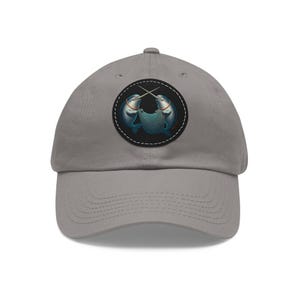 May include: A gray baseball cap featuring a circular patch with two narwhals crossing tusks. The narwhals are blue and white, set against a black background with white stitching. The cap has a curved brim and an adjustable strap.