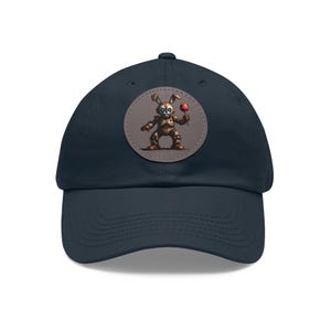 May include: Navy blue baseball cap with a round patch featuring a robotic bunny holding a lollipop. The bunny is brown and has a skull-like face. The lollipop is red. The cap has a curved brim and a fabric strap closure.