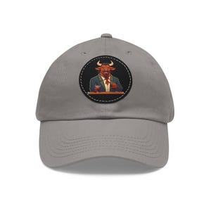 May include: A gray baseball cap featuring a circular patch with a cartoon bull character in a suit, holding a cocktail. The bull has brown fur and is set against a black background. The cap has a curved brim and an adjustable strap.