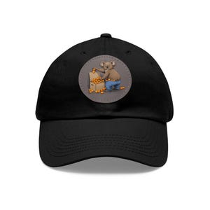 May include: Black baseball cap with a round patch featuring a cartoon koala holding a box of oranges. The patch has a gray background.