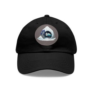 May include: A black baseball cap with a circular patch featuring a white bird standing in a snow-covered cave with blue flowers. The patch has a brown background.
