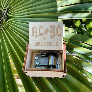 Può includere: Una scatola musicale in legno con un coperchio trasparente. La scatola musicale è aperta e il meccanismo è visibile. La scatola musicale è decorata con le parole "AC/DC" e "hells bells".
