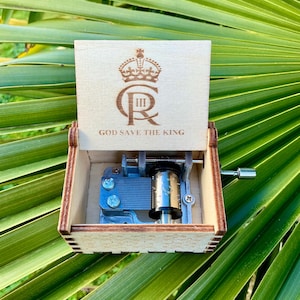 Op de afbeelding: Een houten muziekdoos met een kroon en de tekst "God Save The King" op de deksel. De muziekdoos is open en het mechanisme is zichtbaar.