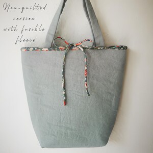 May include: A gray tote bag with a floral patterned trim and tie closure. The bag has long handles and the text "Non-quilted version with fusible fleece" is visible on the upper left side. The bag is made of a soft, textured fabric.