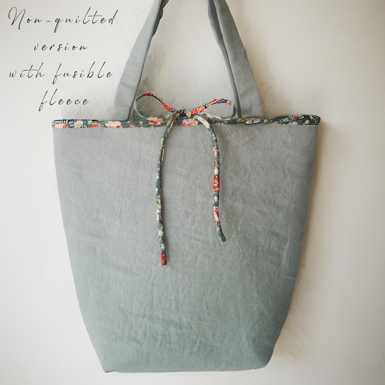 May include: A gray tote bag with a floral patterned trim and tie closure. The bag has long handles and the text "Non-quilted version with fusible fleece" is visible on the upper left side. The bag is made of a soft, textured fabric.