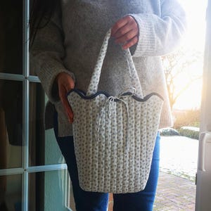 May include: A quilted tote bag with a floral pattern in light blue and white. The bag has a scalloped edge with a dark blue trim and matching handles. A white ribbon is tied at the top. The bag is held by a person.
