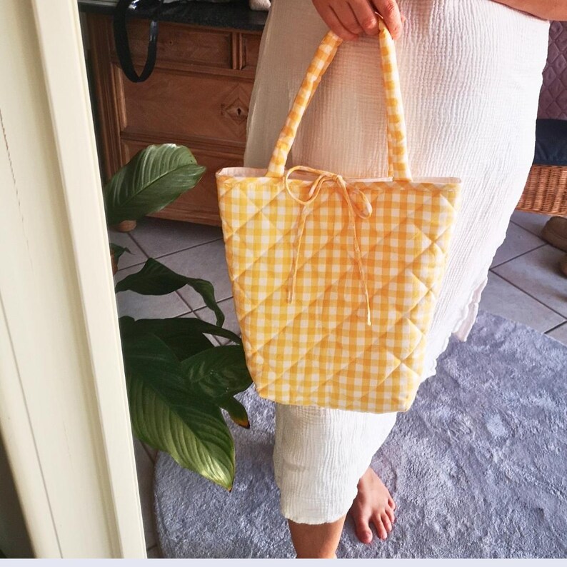 May include: A yellow and white gingham quilted tote bag with matching handles and a bow closure. The bag is held up by a person. The bag is a light yellow color and has a diamond pattern.