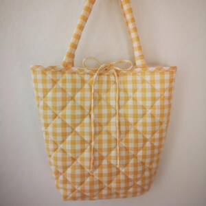 May include: A yellow and white gingham quilted tote bag with matching handles and a bow closure. The bag has a diamond pattern and a scalloped edge. The bag is hanging against a white background.