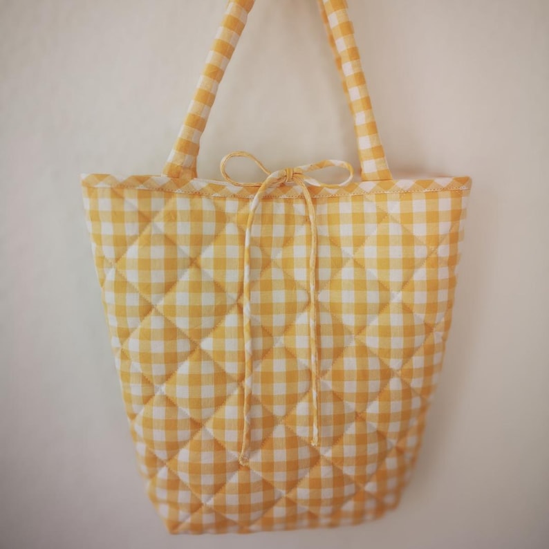 May include: A yellow and white gingham quilted tote bag with matching handles and a bow closure. The bag has a diamond pattern and a scalloped edge. The bag is hanging against a white background.