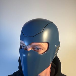 May include: A matte blue helmet with a face shield. The helmet has a sleek, modern design with a black trim and gold accents on the side. The helmet covers the head and face, leaving the eyes exposed.