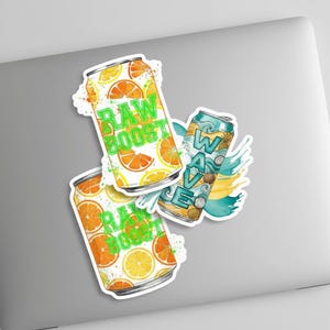 K&ouml;nnte beinhalten: Drei Aufkleber mit farbenfrohen Illustrationen von Getr&auml;nkedosen. Zwei Dosen sind mit "RAW BOOST" mit Orangen- und Zitronenscheiben beschriftet. Die dritte Dose ist mit "WAVE" mit tropischem Design und t&uuml;rkisfarbenen Akzenten beschriftet.