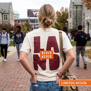 May include: A cream-colored short-sleeve t-shirt featuring large dark red letters spelling LAND on the back. An orange rectangular patch is centered over the letters with the white text NEVER NEVER printed on it. The shirt is worn with blue jeans.