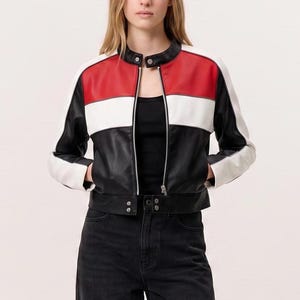Puede incluir: Una chaqueta de bloques de color con un panel superior rojo, una franja central blanca y una sección inferior negra. La chaqueta tiene cuello alto, una cremallera central plateada, mangas largas con puños blancos y dos botones de presión.