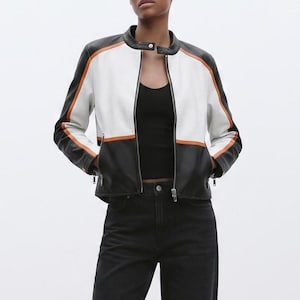 Può includere: Una giacca a blocchi di colore con un design bianco e nero e sottili strisce orizzontali arancioni. La giacca presenta un colletto alla coreana con chiusura a bottone automatico, una zip frontale e due tasche con cerniera sul davanti.