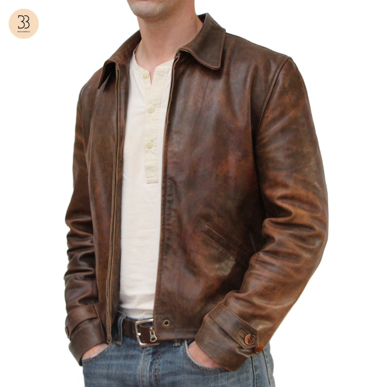 May include: A brown leather jacket with a collared neckline and a zipper closure. The jacket has a distressed appearance and features a buttoned cuff on the sleeves. The person is wearing a white shirt and blue jeans.