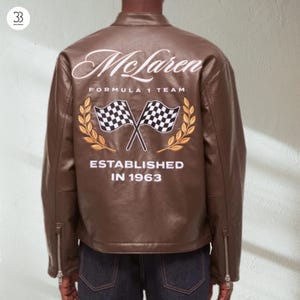 May include: A brown leather jacket featuring the McLaren Formula 1 Team logo embroidered on the back. The design includes crossed checkered flags, laurel leaves, and the text "ESTABLISHED IN 1963". The jacket has a stand-up collar and zippered pockets.