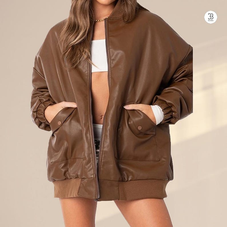 May include: A brown faux leather bomber jacket with a zip-up front and two front pockets. The jacket has a ribbed hem and cuffs. The model is wearing a white top underneath. The jacket is a relaxed fit.