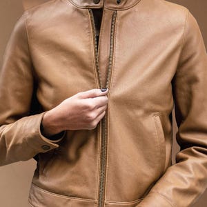 Puede incluir: Chaqueta de cuero color canela con cuello alto y cremallera completa. La chaqueta tiene dos bolsillos delanteros y detalles de botones en los pu&ntilde;os. La persona est&aacute; subiendo la cremallera.