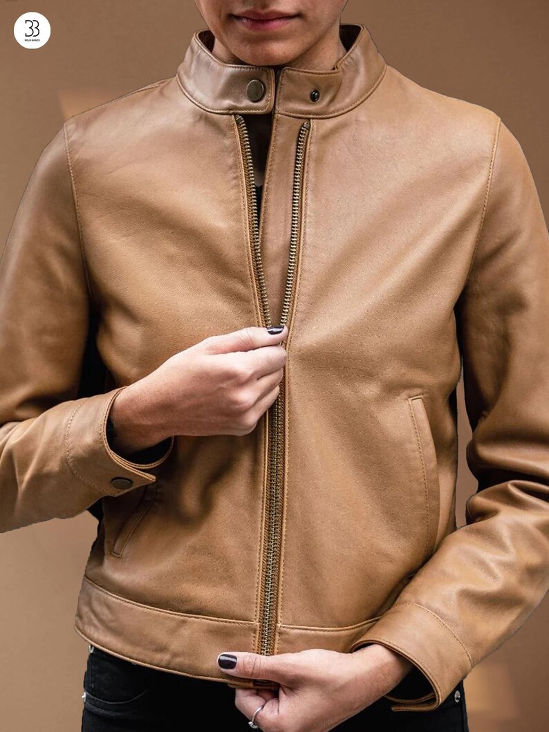Puede incluir: Chaqueta de cuero color canela con cuello alto y cremallera completa. La chaqueta tiene dos bolsillos delanteros y detalles de botones en los pu&ntilde;os. La persona est&aacute; subiendo la cremallera.