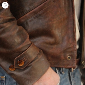 May include: Close-up of a brown leather jacket with a distressed finish. The jacket features a buttoned cuff detail with two brown buttons. The leather has a worn appearance, and the jacket is partially zipped.