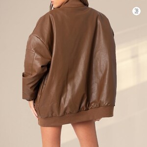 May include: A brown faux leather bomber jacket with a high collar and ribbed cuffs and hem. The jacket has a relaxed fit and a smooth, shiny texture. The back view shows the jacket's full shape and design.