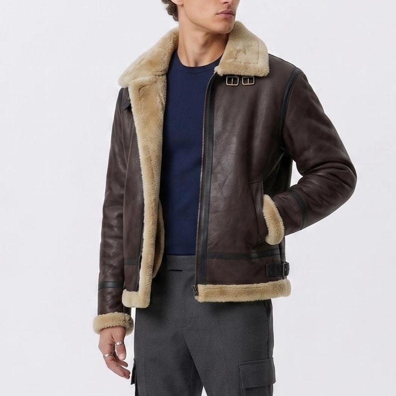 May include: A dark brown jacket with a thick beige shearling lining and collar. The jacket features a front zipper closure, two metal buckles at the neck, and black leather trim accents along the seams, cuffs, and hemline for a structured look.