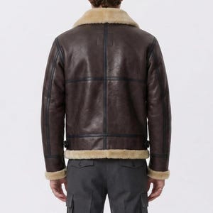 May include: A dark brown leather jacket featuring a tan shearling collar and trim at the cuffs and hem. The jacket has visible dark seam detailing, a structured back panel, and adjustable side tabs at the waist, worn over dark gray trousers.