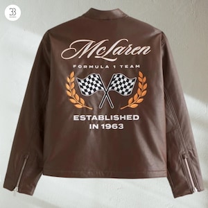 May include: A brown leather jacket featuring the McLaren Formula 1 Team logo on the back. The logo includes the word "McLaren" in white script, crossed checkered flags, and the text "ESTABLISHED IN 1963". The jacket has zip details on the sleeves.