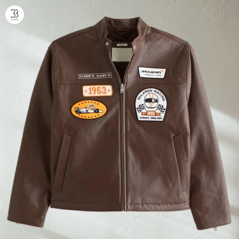 May include: A brown leather jacket with a zip-up front and a stand-up collar. The jacket features several patches, including a "1963" patch and a "McLaren Racing" patch. The jacket has two front pockets and a small tag near the collar.