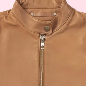 Puede incluir: Primer plano de una chaqueta de cuero color canela con cuello alzado y cremallera completa. La chaqueta tiene un dise&ntilde;o cl&aacute;sico con cierre de bot&oacute;n en el cuello y un acabado liso y pulido. La chaqueta es de color neutro.