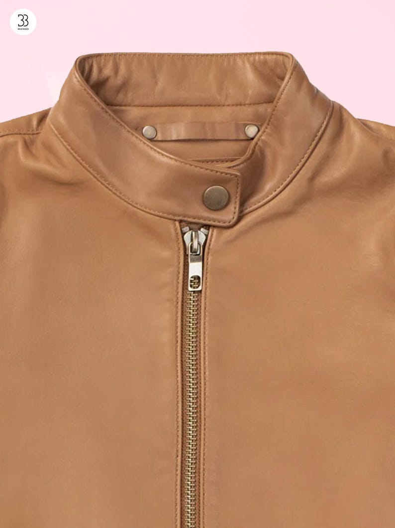 Puede incluir: Primer plano de una chaqueta de cuero color canela con cuello alzado y cremallera completa. La chaqueta tiene un dise&ntilde;o cl&aacute;sico con cierre de bot&oacute;n en el cuello y un acabado liso y pulido. La chaqueta es de color neutro.