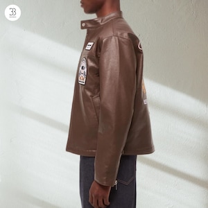 May include: A brown leather jacket with a stand-up collar and several patches. The jacket has a classic design with a button closure and a tailored fit. The patches feature various designs and text. The jacket is paired with dark denim jeans.