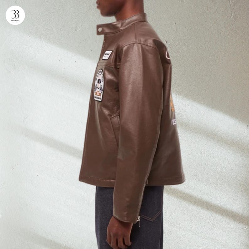 May include: A brown leather jacket with a stand-up collar and several patches. The jacket has a classic design with a button closure and a tailored fit. The patches feature various designs and text. The jacket is paired with dark denim jeans.