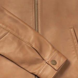 Puede incluir: Primer plano de una chaqueta de cuero color canela. La chaqueta tiene una cremallera dorada, un pu&ntilde;o abotonado y un peque&ntilde;o bolsillo. El cuero tiene una textura suave y un color c&aacute;lido y neutro.