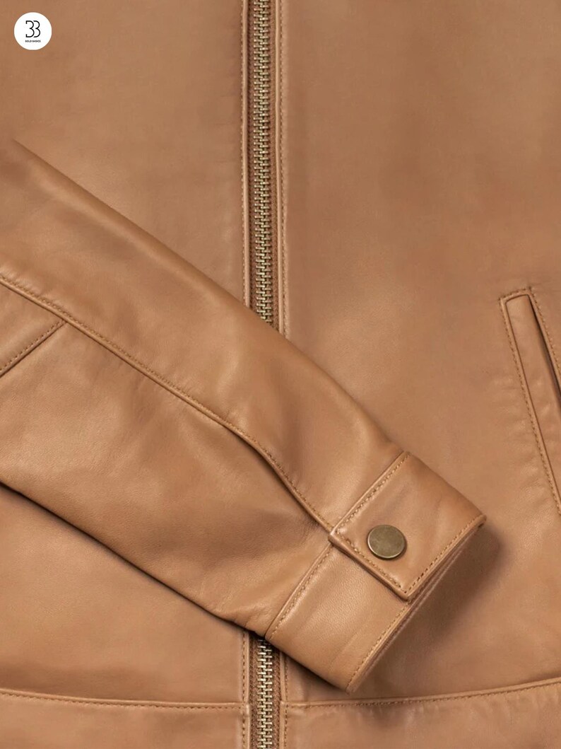 Puede incluir: Primer plano de una chaqueta de cuero color canela. La chaqueta tiene una cremallera dorada, un pu&ntilde;o abotonado y un peque&ntilde;o bolsillo. El cuero tiene una textura suave y un color c&aacute;lido y neutro.