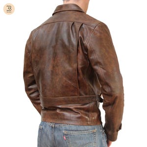 May include: Brown leather jacket with a classic design. The jacket features a collar, a belt at the waist, and a distressed finish. The back of the jacket has a unique pleated detail. The model is wearing blue jeans.
