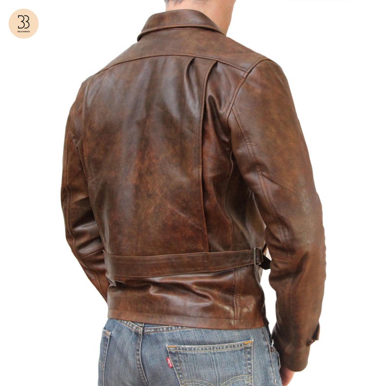 May include: Brown leather jacket with a classic design. The jacket features a collar, a belt at the waist, and a distressed finish. The back of the jacket has a unique pleated detail. The model is wearing blue jeans.