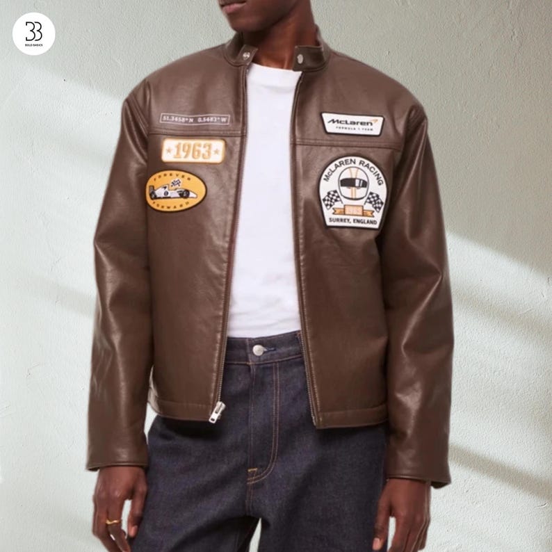 May include: Brown leather jacket with a stand-up collar and various patches. Patches include "1963" and "McLaren Racing". The jacket features a zip closure and is worn with a white t-shirt and dark blue jeans.