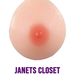 May include: A prosthetic breast form in a natural skin tone, featuring a pink areola. The image includes the text "JANETS CLOSET DETROIT, MICHIGAN" in purple. The form is designed to mimic the appearance of a natural breast.