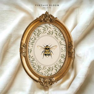May include: An oval, gold-colored ornate frame surrounds a print featuring a watercolor bee illustration. The bee is centered on a cream-colored background with a floral pattern. The text "VINTAGE BLOOM PRINTS" is at the top.