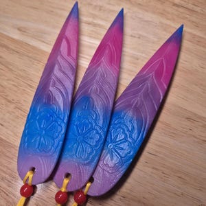 Könnte beinhalten: Drei dekorative, längliche Anhänger mit floralem Design, bemalt in einem Farbverlauf aus Blau, Lila und Rosa. Jeder Anhänger hat eine gelbe Schnur mit einer roten Perle. Die Anhänger werden auf einer Holzoberfläche präsentiert.