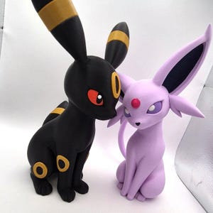 Könnte beinhalten: Zwei stehende Tierfiguren. Eine ist ein schwarzes Wesen mit gelben Ringen, roten Augen und langen Ohren. Die andere ist ein hellviolettes Wesen mit großen spitzen Ohren, einem dunklen Innenohr und einem kleinen roten runden Edelstein auf der Stirn.