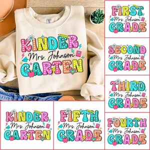 Puede incluir: Sudadera crema con el texto "Kinder Mrs. Johnson Garten" en letras de burbujas coloridas. La imagen también muestra diseños de niveles de grado de primero a quinto grado. Incluye un lápiz, una manzana y un rayo.