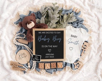 以下が含まれることがあります： 「We are excited to say Baby Boy is on the way. Arriving July 2024.」と書かれた黒い文字盤。白いロンパースには「Baby Smith」と小さなハートが書かれている。BABYと綴られた木製のブロックと、その他のベビー用品の装飾品。