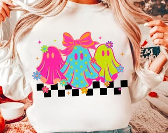 May include: White sweatshirt featuring a colorful Halloween design. The graphic includes three cartoon ghosts in pink, blue, and green, adorned with bows, flowers, and stars. A black and white checkered pattern is at the bottom of the design.