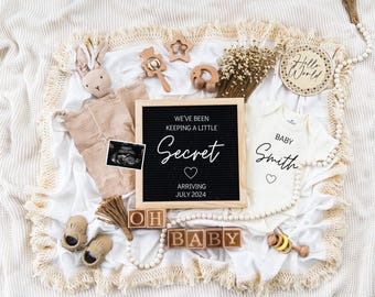 May include: A white blanket with a black letter board that reads "We've been keeping a little Secret Arriving July 2024". There is a white onesie with the text "Baby Smith" on it. There are also wooden blocks that spell out "Oh Baby" and other baby items.