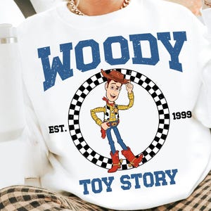 Puede incluir: Sudadera blanca con un gr&aacute;fico de Woody de Toy Story. El dise&ntilde;o incluye la palabra "WOODY" en letras azules en bloque sobre una imagen circular del personaje. Las palabras "EST. 1999" y "TOY STORY" tambi&eacute;n est&aacute;n impresas en la sudadera.